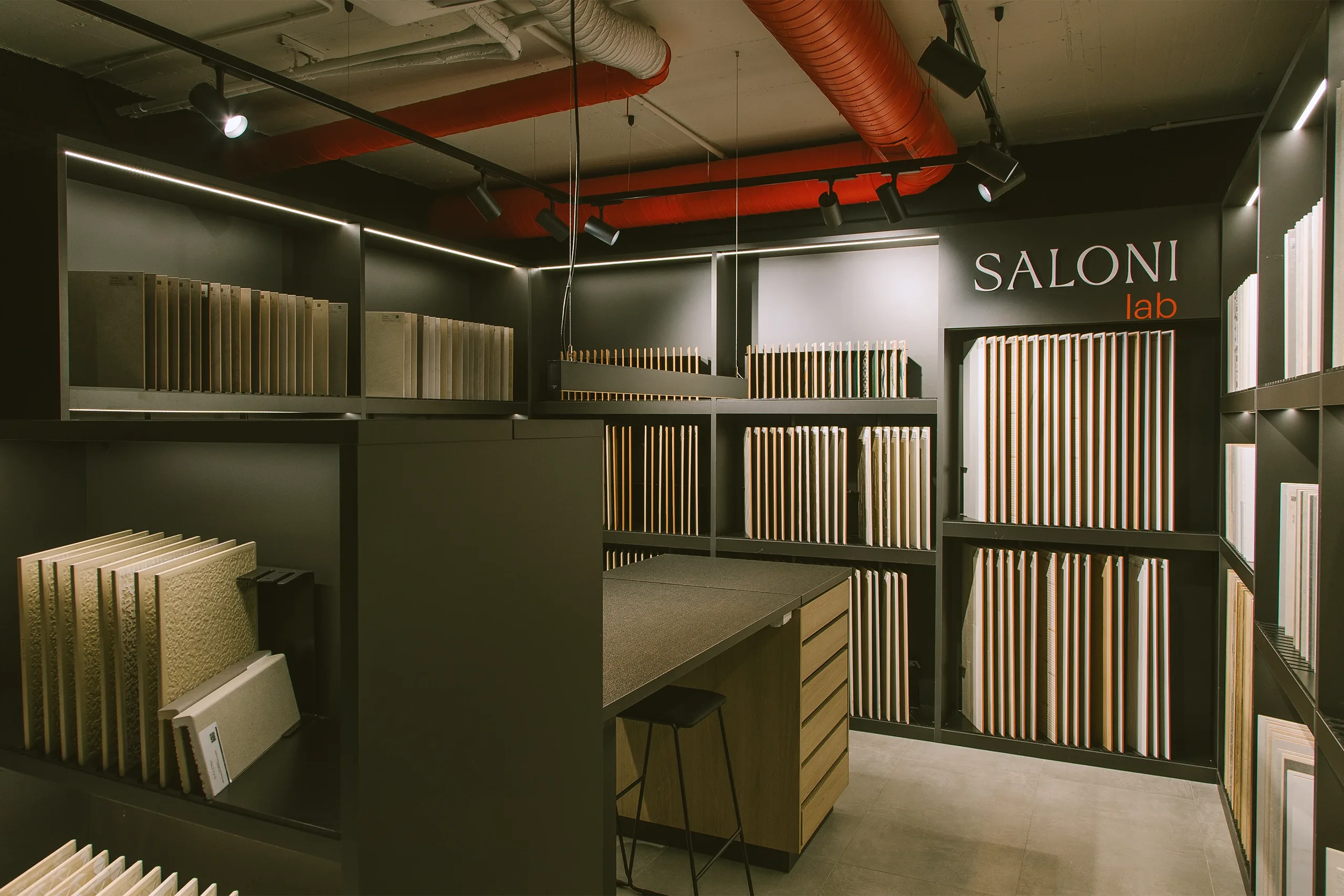 Zona SALONI Lab del showroom Madrid con estanterías negras de muestrario cerámico, conductos rojos vistos e iluminación lineal