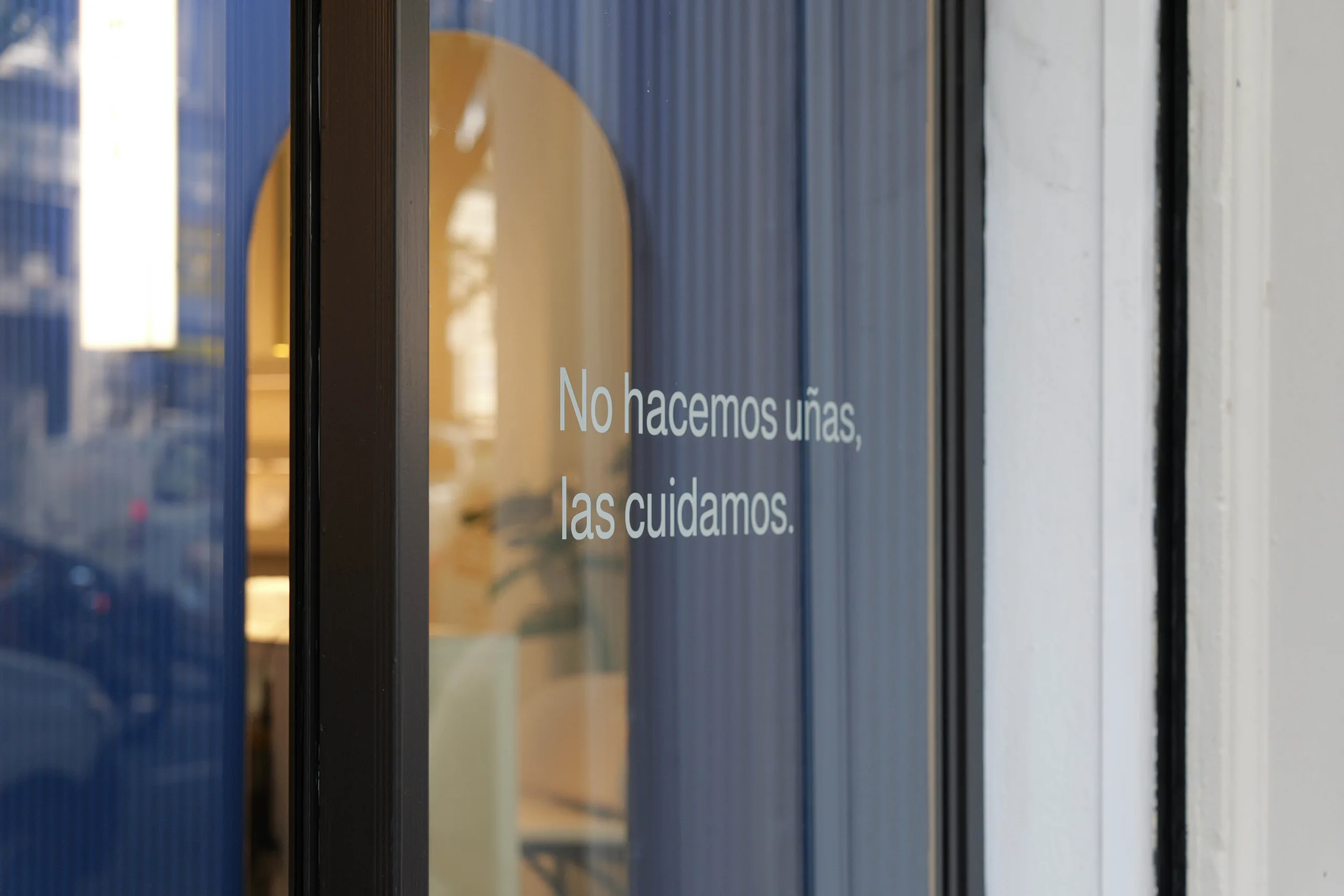 Detalle del escaparate de Arpías Avenida Gaudí (Eixample, Barcelona) con vinilo "No hacemos uñas, las cuidamos", panel acanalado azul y arco interior visible al fondo