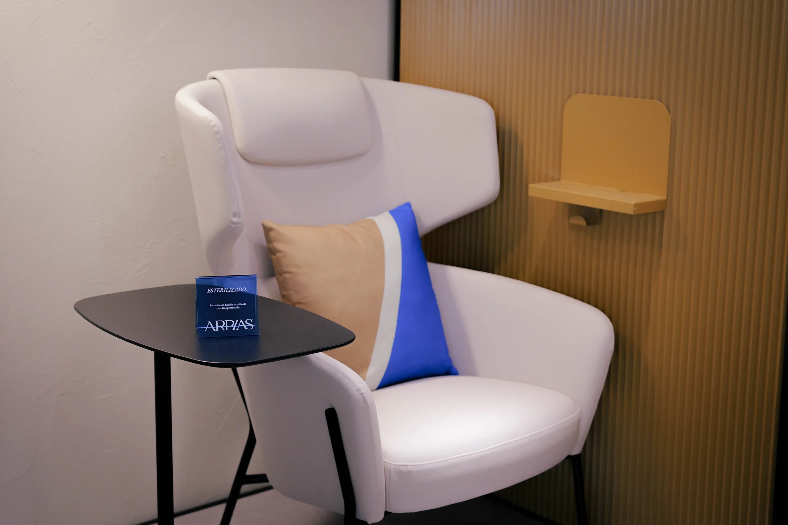 Zona La Butaca de pedicura en Arpías Avenida Gaudí (Eixample, Barcelona) con butaca tapizada blanca, cojín azul y beige, mesa auxiliar y panel acanalado de roble