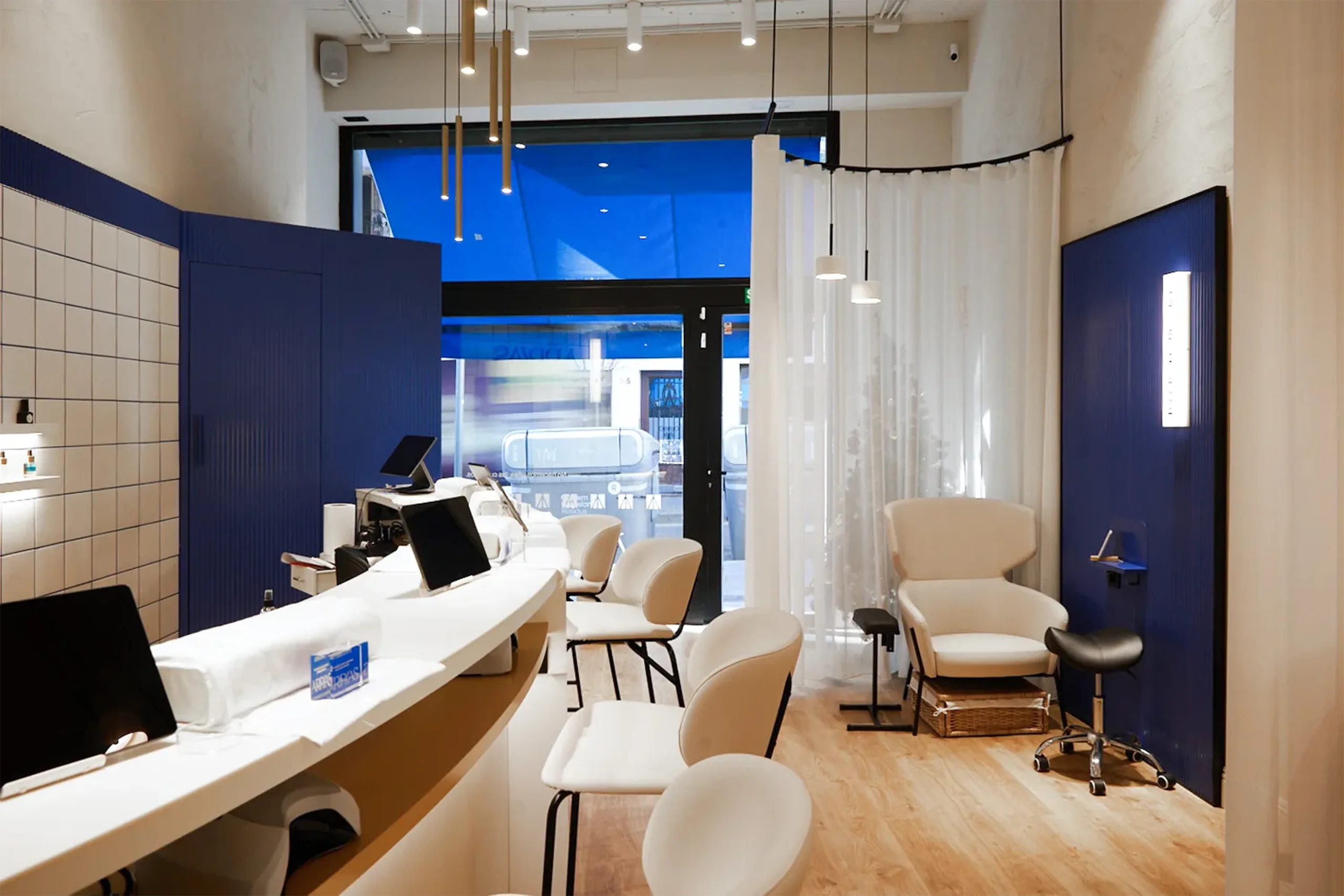 Vista interior del salón Arpías en Torrent de l'Olla (Gràcia, Barcelona) con barra de manicura, sillón de pedicura, cortinas translúcidas y paneles acanalados azules