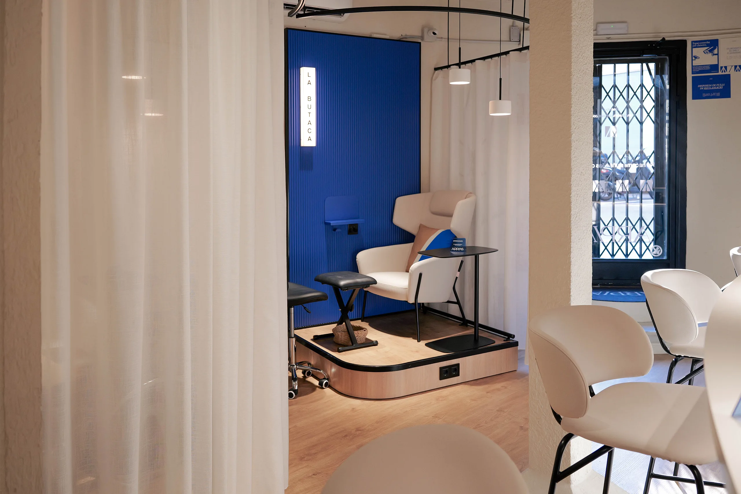 Sala privada La Butaca en Arpías Putxet (calle Rubinstein, Barcelona) con butaca tapizada blanca, panel acanalado azul con rótulo retroiluminado, cortinas blancas translúcidas y tarima de madera