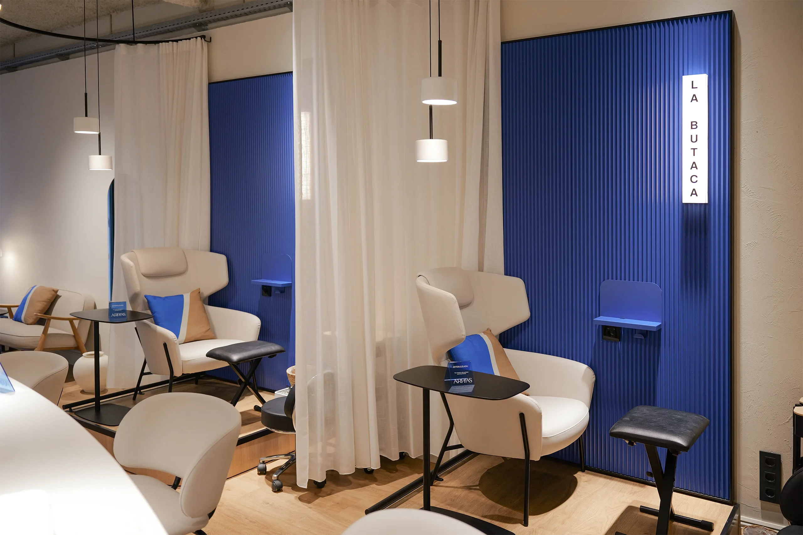 Zona La Butaca del salón Arpías en Granada del Penedés (Sant Gervasi, Barcelona) con sillones de pedicura, paneles acanalados azules, cortinas translúcidas y techo industrial