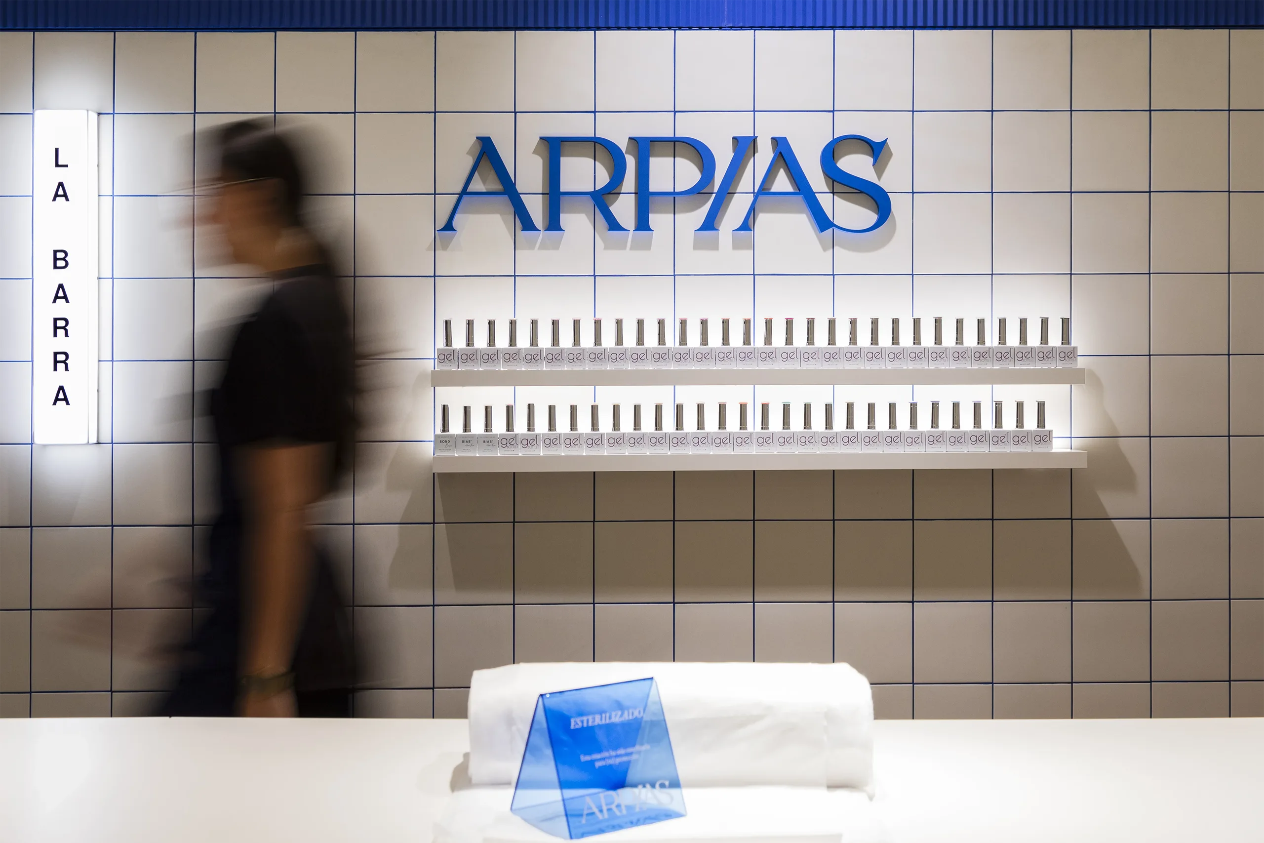 Pared de La Barra en Arpías Madrazo (Sant Gervasi, Barcelona) con alicatado cerámico blanco con junta azul, rótulo de marca y exposición lineal de esmaltes en gel