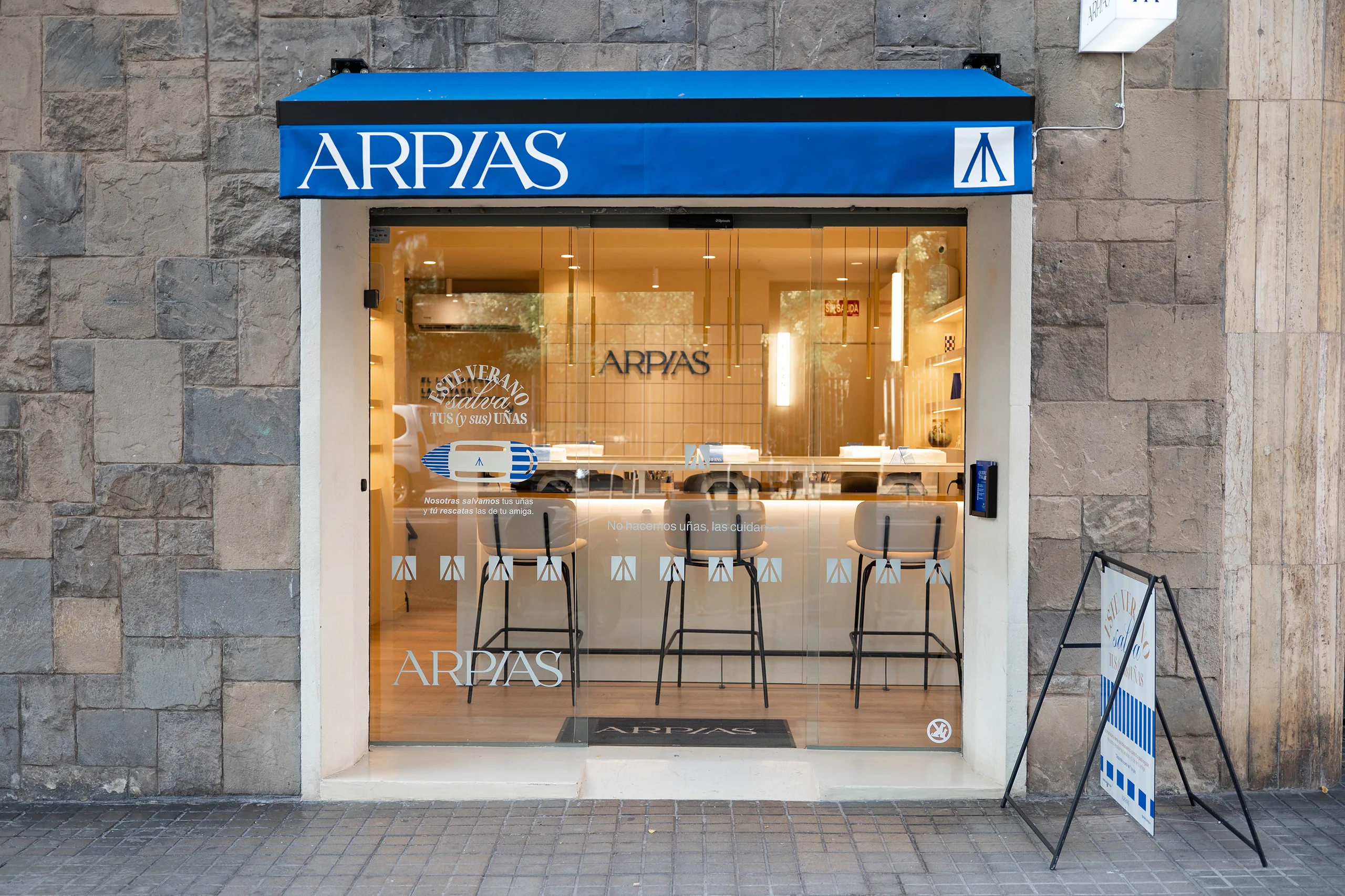Fachada de Arpías en Turó Park (Sant Gervasi, Barcelona) con muro de piedra original del edificio, toldo azul, rótulo perpendicular y escaparate del salón