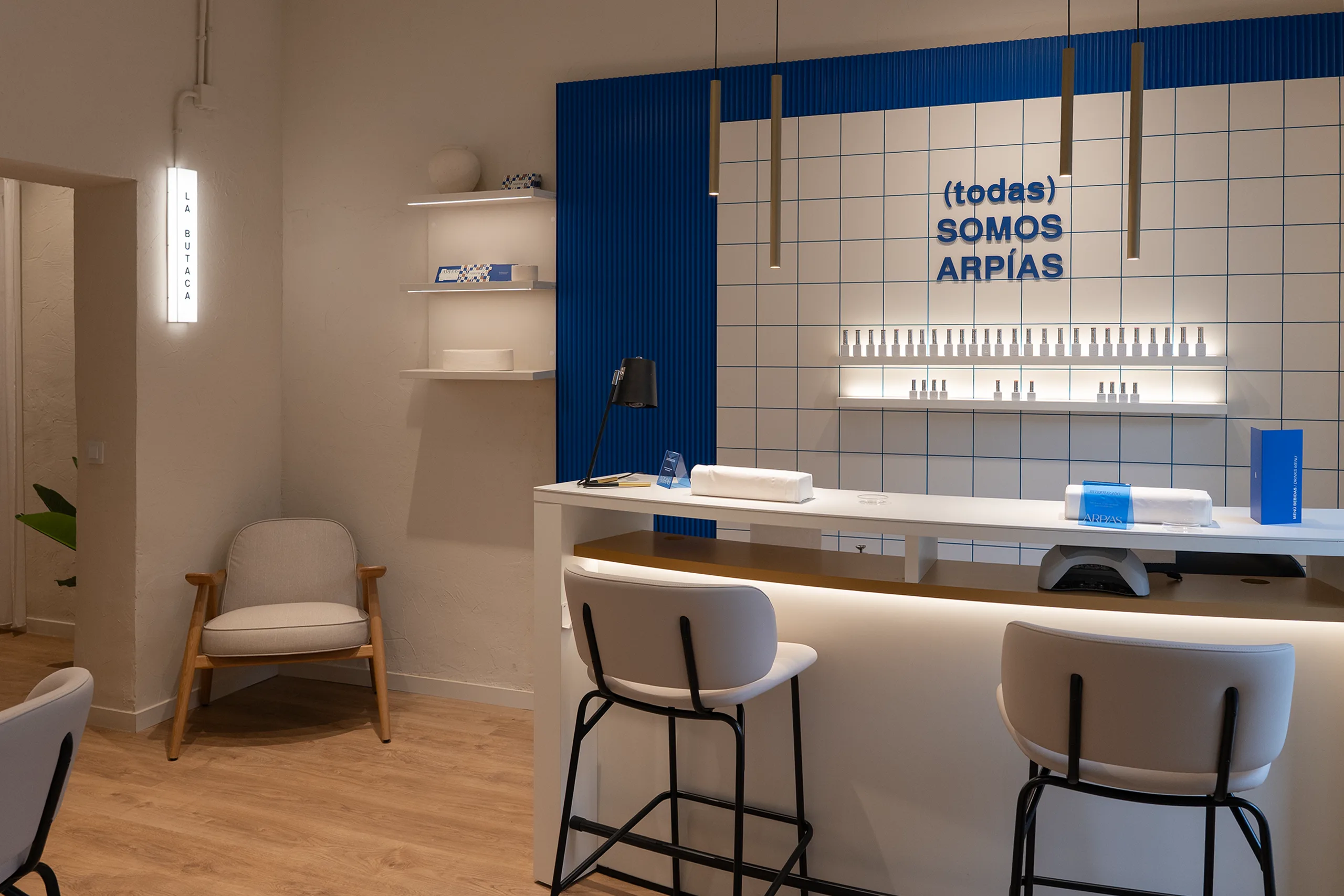 La Barra en Arpías Atocha (Centro, Madrid) con barra alargada de manicura, rótulo "(todas) somos Arpías" sobre alicatado cerámico blanco con junta azul, butaca de espera lateral y rótulo retroiluminado "LA BUTACA"