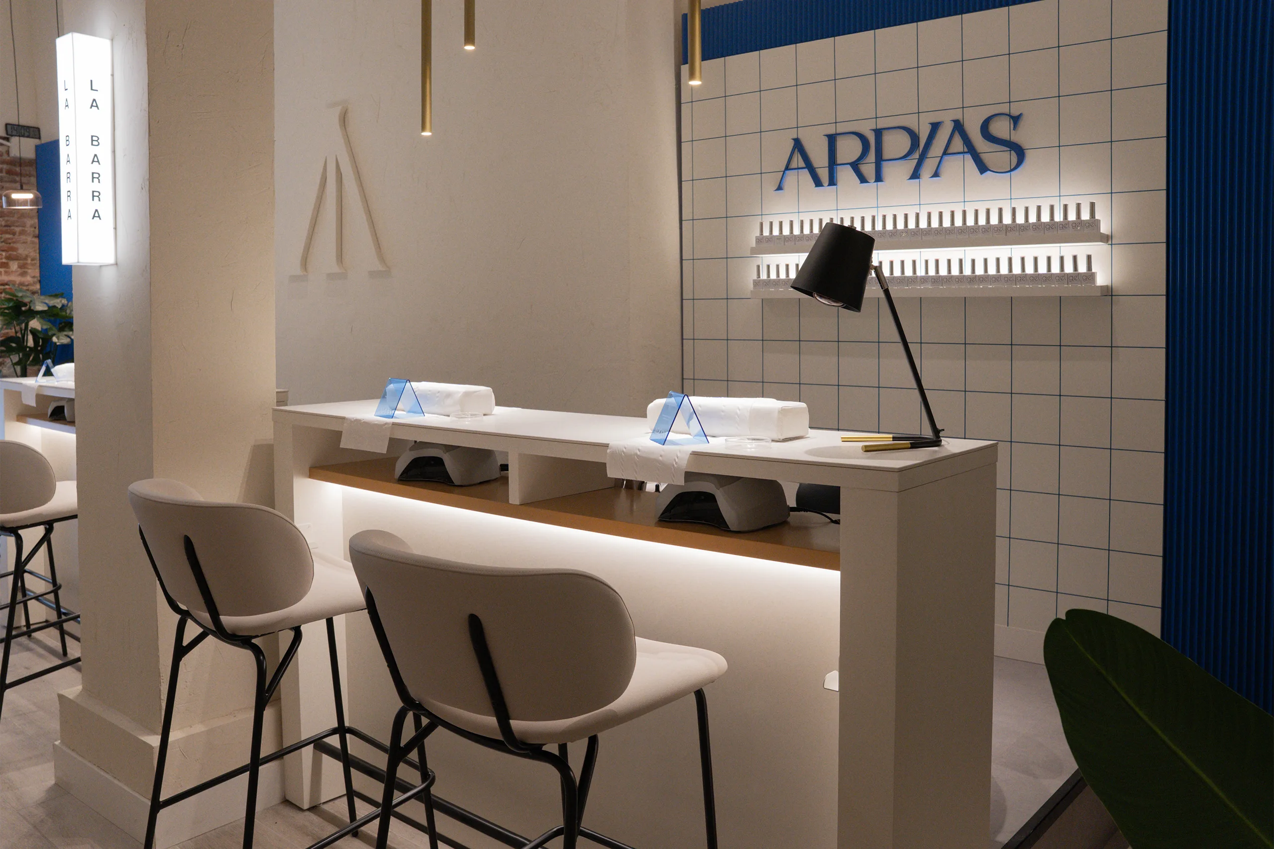 Detalle de un puesto de La Barra en Arpías Libertad (Chueca, Madrid) con alicatado cerámico blanco con junta azul, rótulo retroiluminado, exposición de esmaltes en gel y lámpara de mesa