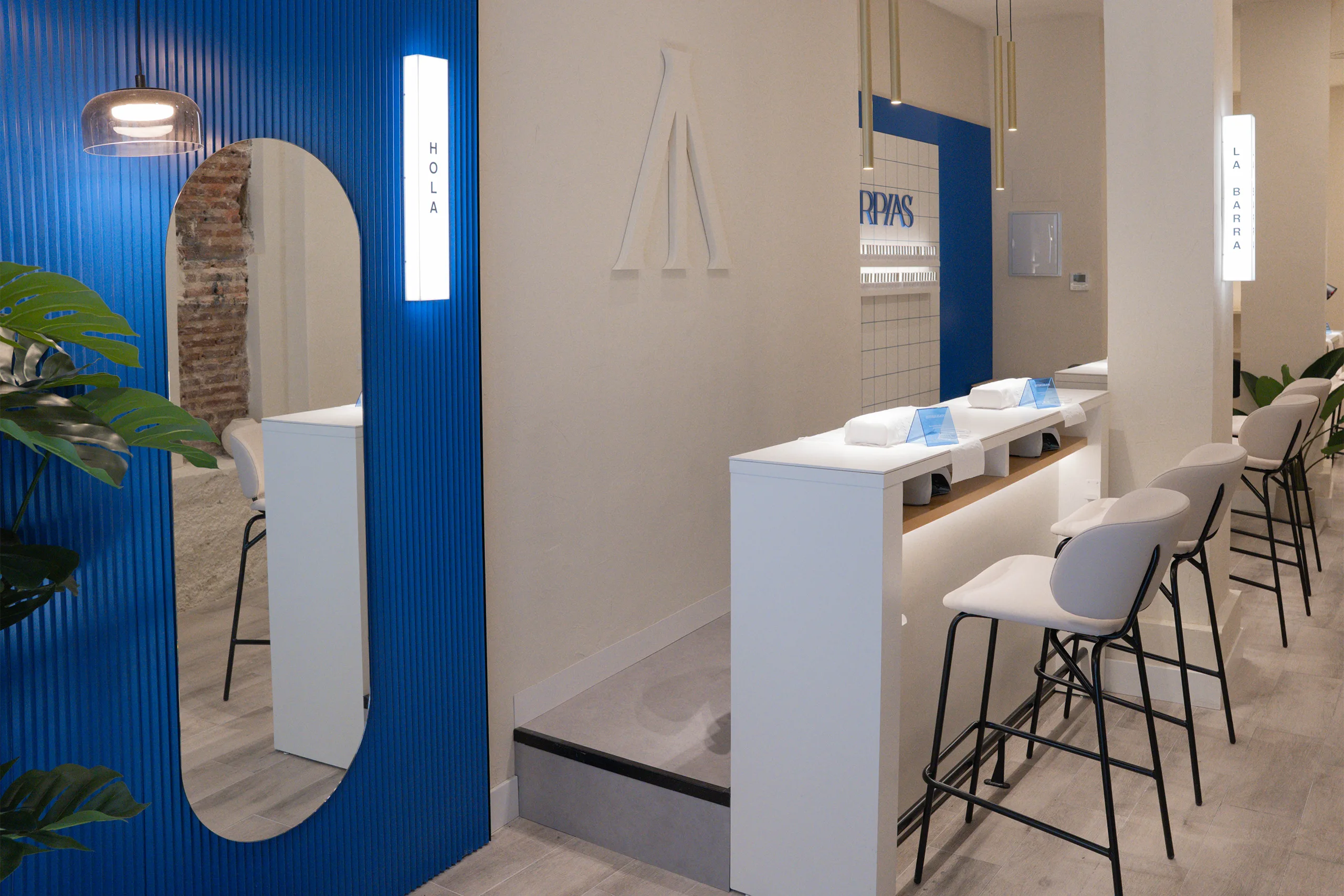 Zona de bienvenida en Arpías Libertad (Chueca, Madrid) con paneles acanalados azules, rótulo retroiluminado "HOLA", espejo de cuerpo entero y lámpara colgante