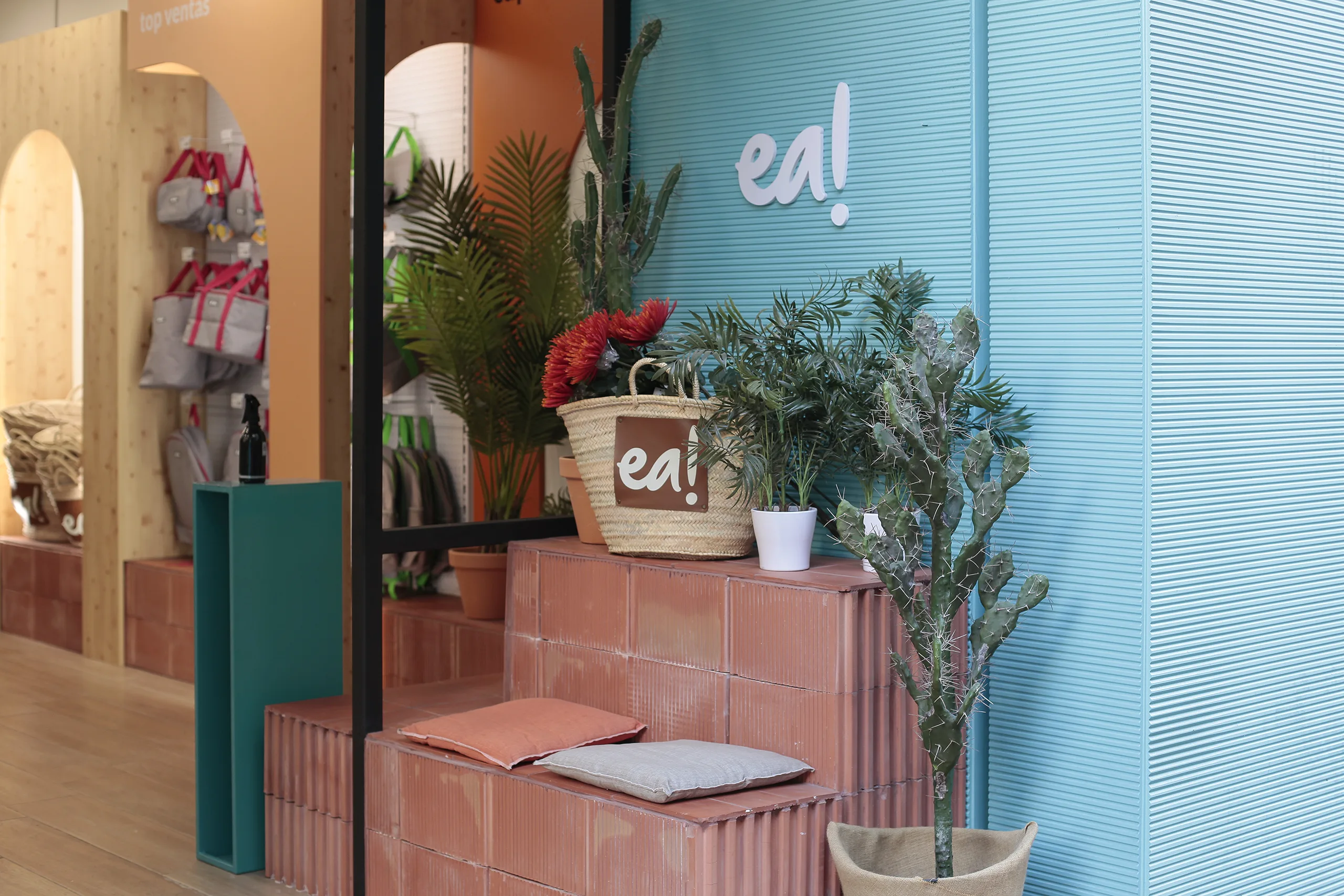 Detalle del exterior de caprichea! Almería con muro azul turquesa rayado, rótulo "ea!" en blanco, capazo con flores y cactus sobre escalonado de ladrillo en arcilla