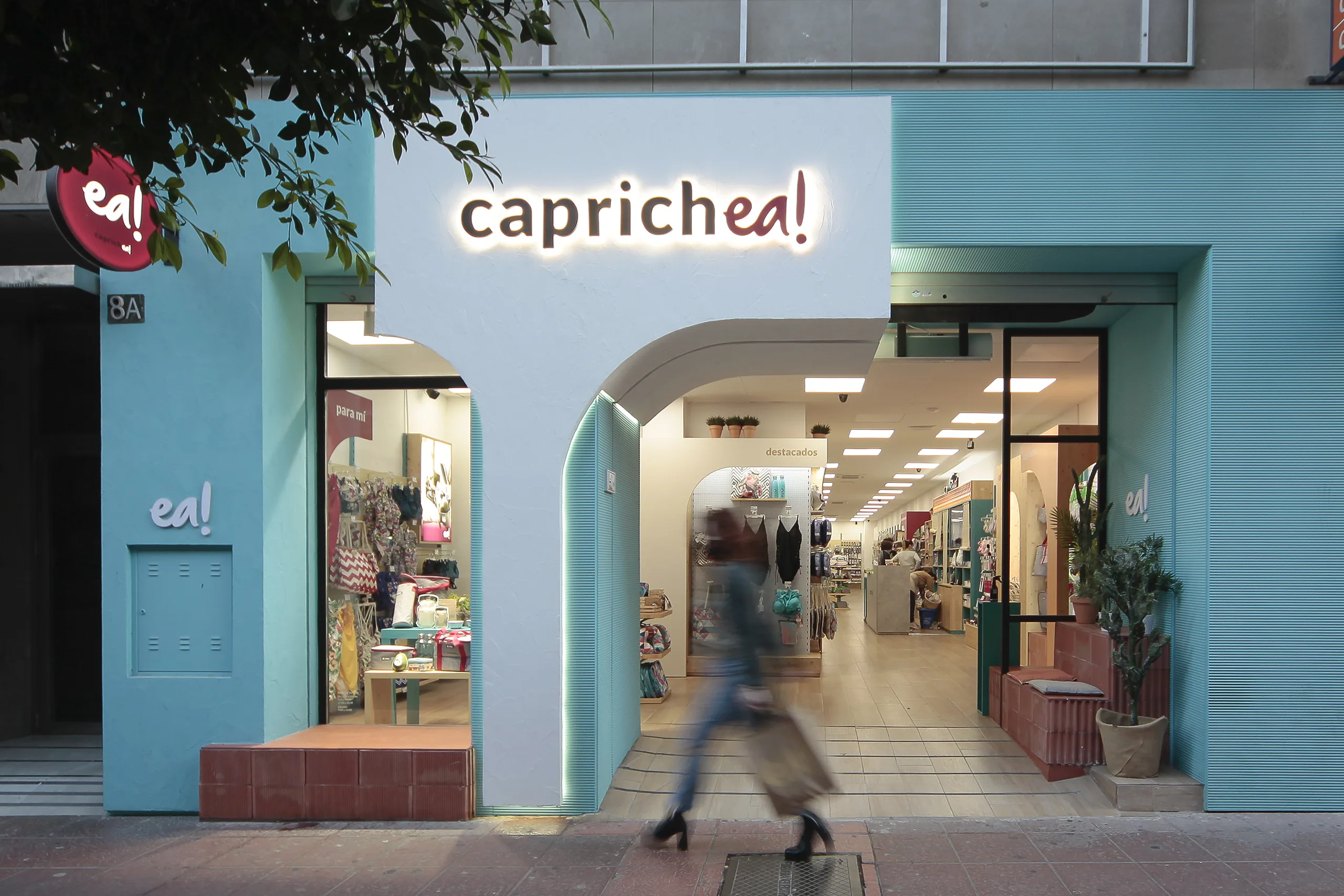 Fachada de la tienda Caprichea! en Almería con arco mediterráneo blanco, fondo azul turquesa, rótulo retroiluminado "caprichea!" y zócalo en arcilla