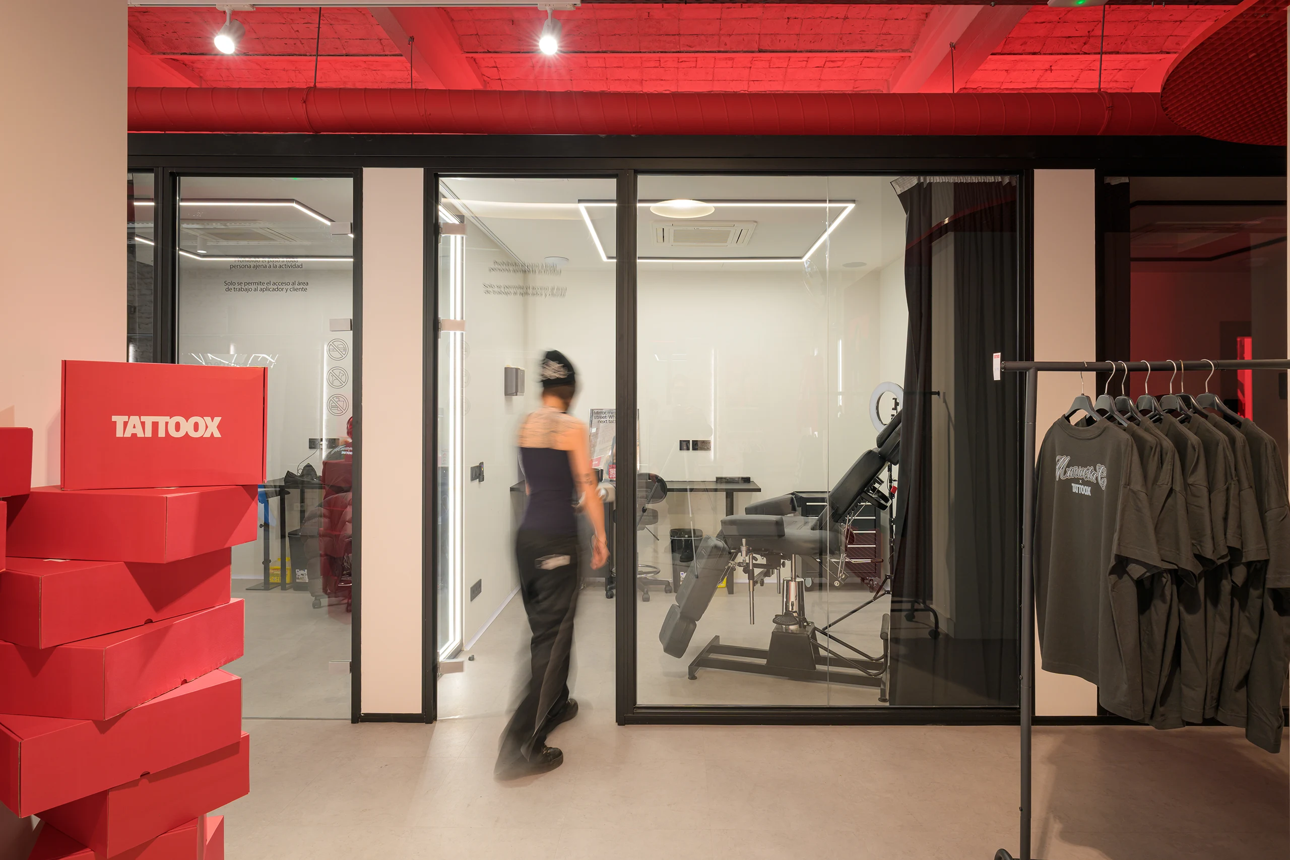 Cabina de tatuaje acristalada en Tattoox Club Barcelona con techo de ladrillo visto teñido en rojo, sillón profesional y zona de retail