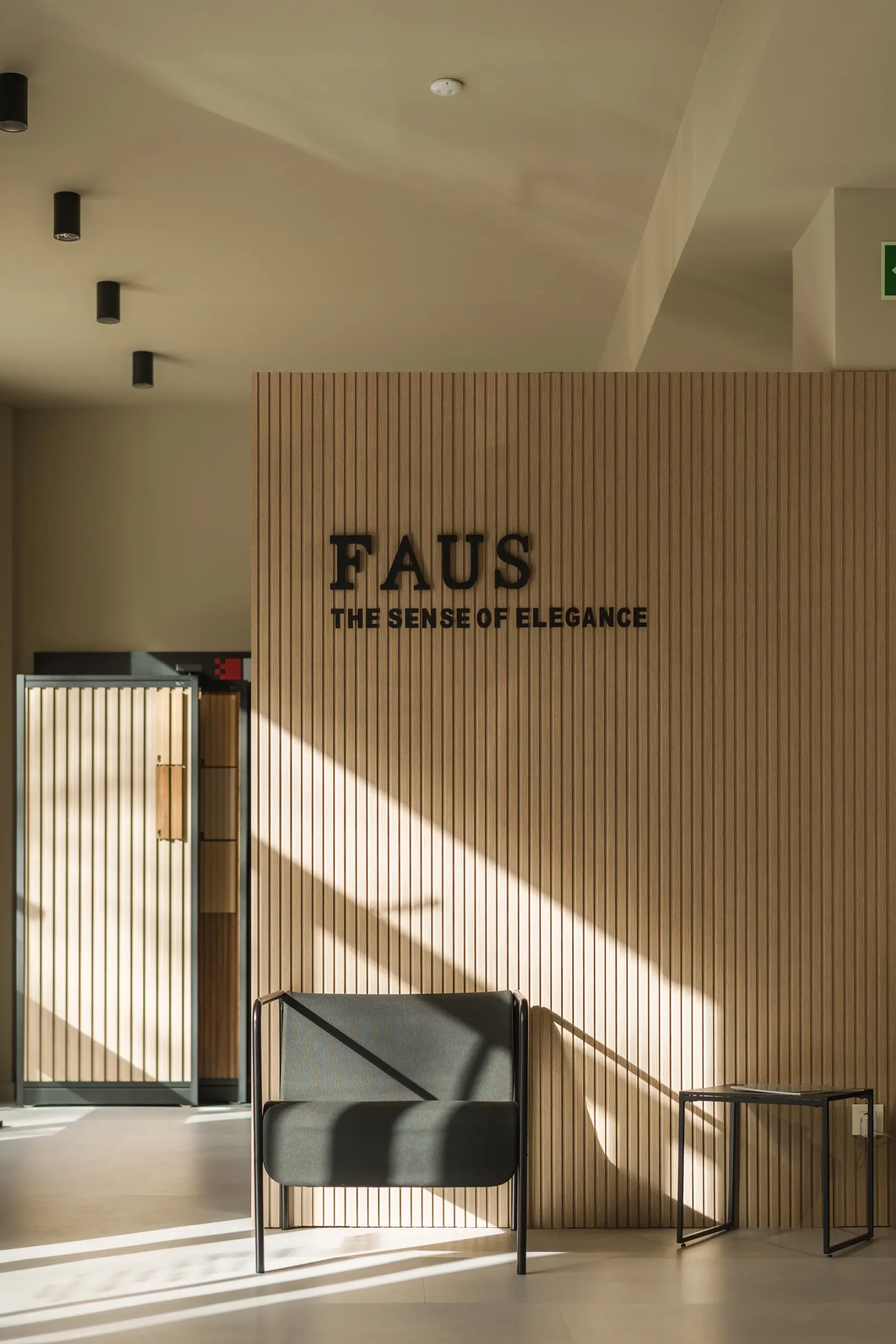 Muro de lamas de madera con logotipo FAUS The Sense of Elegance, butaca negra y luz natural rasante