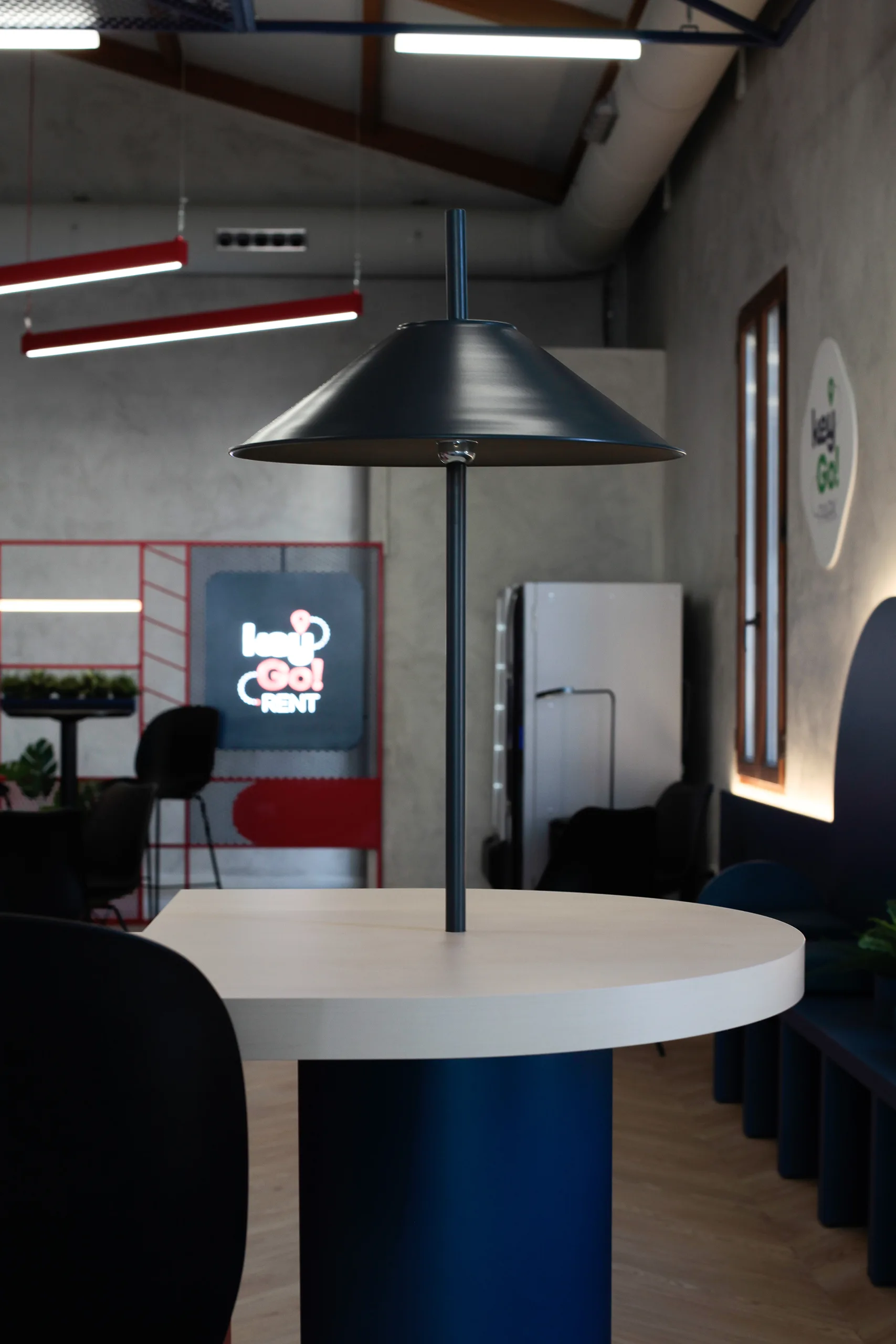 Detalle de mesa alta con tablero claro y base azul petróleo en Key Go Rent! Alicante, con lámpara negra y rótulo de neón desenfocado