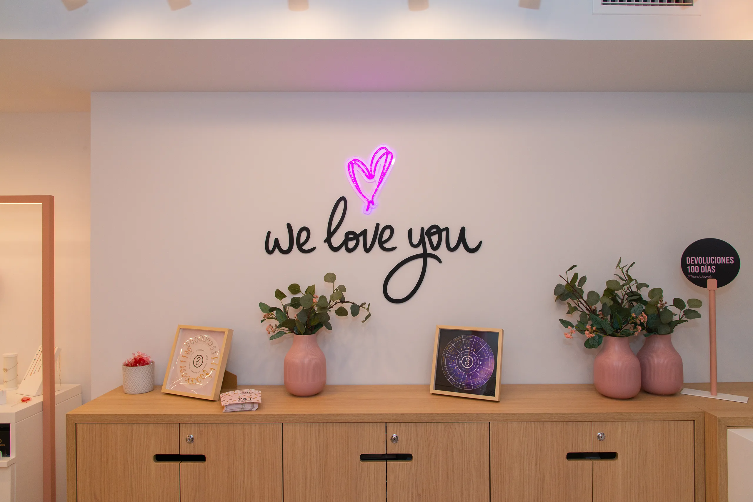 Pared blanca de tienda Singularu con rótulo we love you y corazón de neón rosa sobre mueble de madera con jarrones